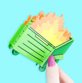 A hand with purple nails holds a novelty pin resembling a green dumpster fire, suggesting humor or chaos