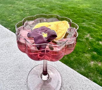 Decorative glass bowl with floral-shaped rim, holding a pink liquid and floating colorful flower petals, placed outdoors on a concrete surface