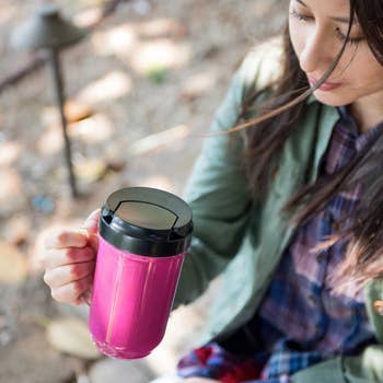 Person holding a travel mug outdoors, wearing a plaid shirt under a jacket, suggesting comfort and style for on-the-go beverage needs