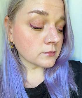 Person with lavender hair and nose ring, wearing a black top, showcasing shimmery eye makeup and gold hoop earrings
