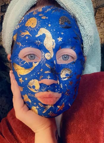 Person wearing a towel-turban and celestial-themed sheet mask with gold designs, posing with a hand on cheek
