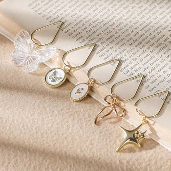 Five gold bookmarks with charms: a butterfly, bee, flower, bow, and star, lay on an open book, suggesting elegant reading accessories