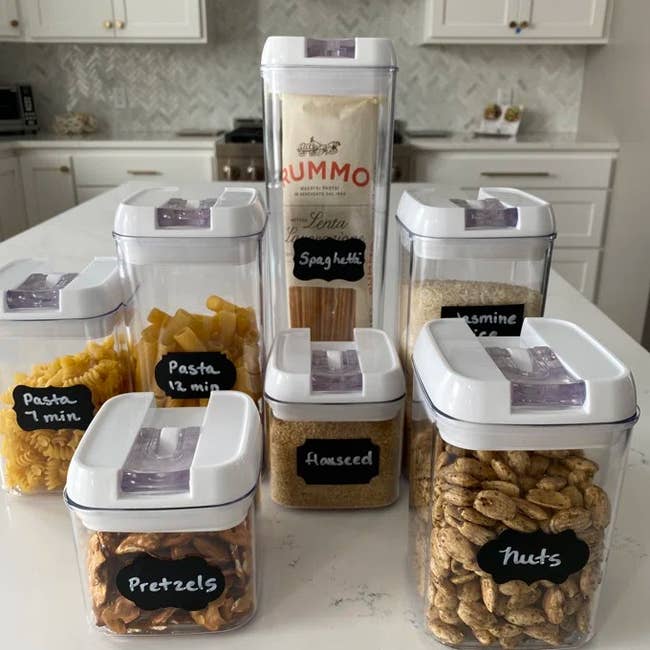 Clear kitchen containers labeled for pasta, flaxseed, pretzels, and nuts on a countertop, showcasing organized food storage options