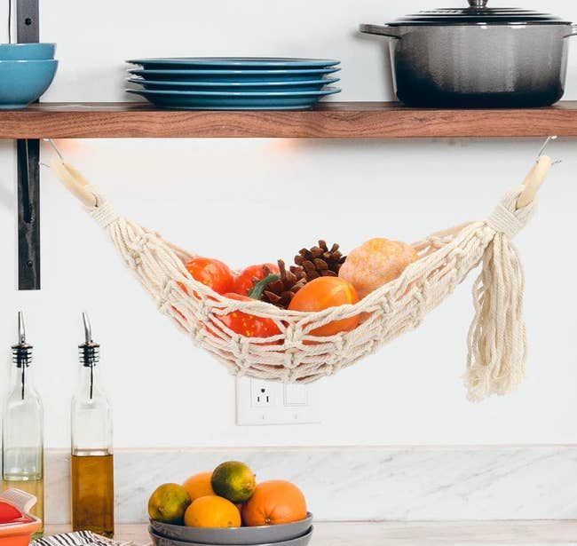 Hanging woven produce hammock under a kitchen shelf, filled with fruits and vegetables. Icons highlight features: 100% cotton, easy to wash, hand woven