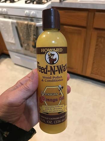 A hand holds a bottle of Howard Feed-N-Wax Wood Polish & Conditioner