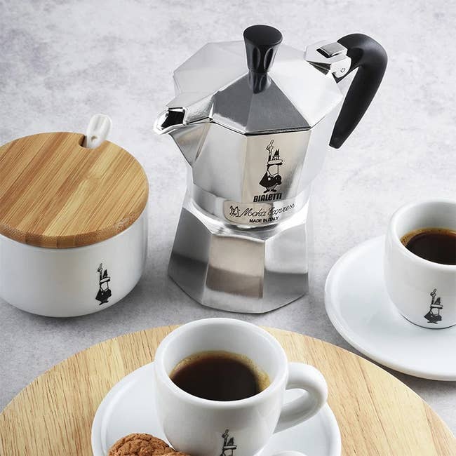 Espresso maker set with a metal coffee pot and two cups on saucers, all branded, alongside a sugar bowl with a wooden lid on a table