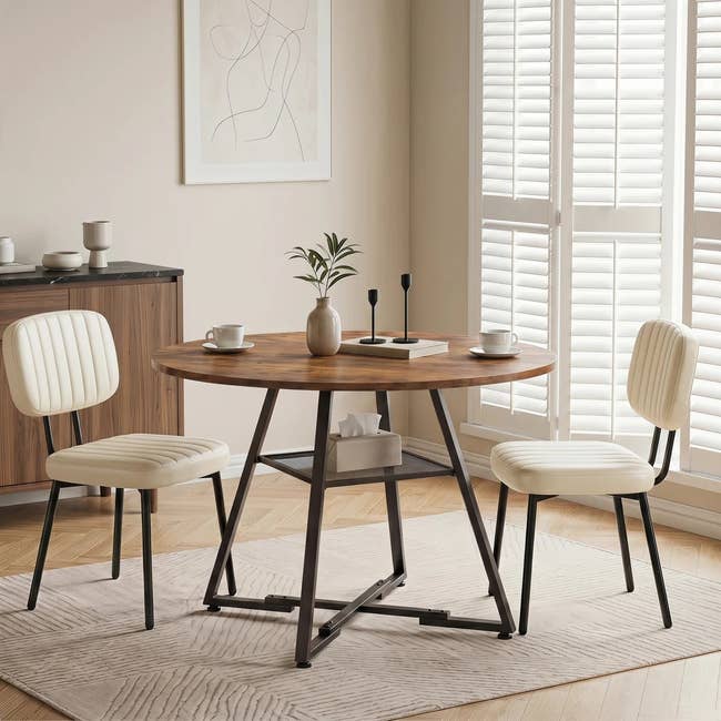 Dining area with a round wooden table, two cream chairs, and minimalist decor. Natural light filters through shutters, creating a cozy atmosphere