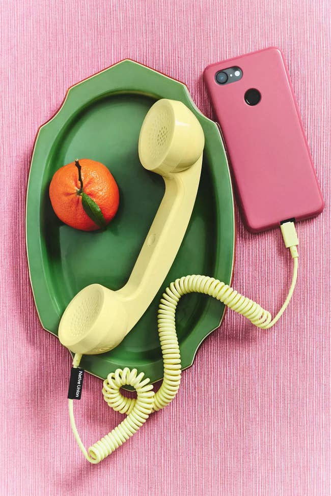 Retro yellow corded phone handset connected to a modern pink smartphone placed on a green tray with an orange fruit
