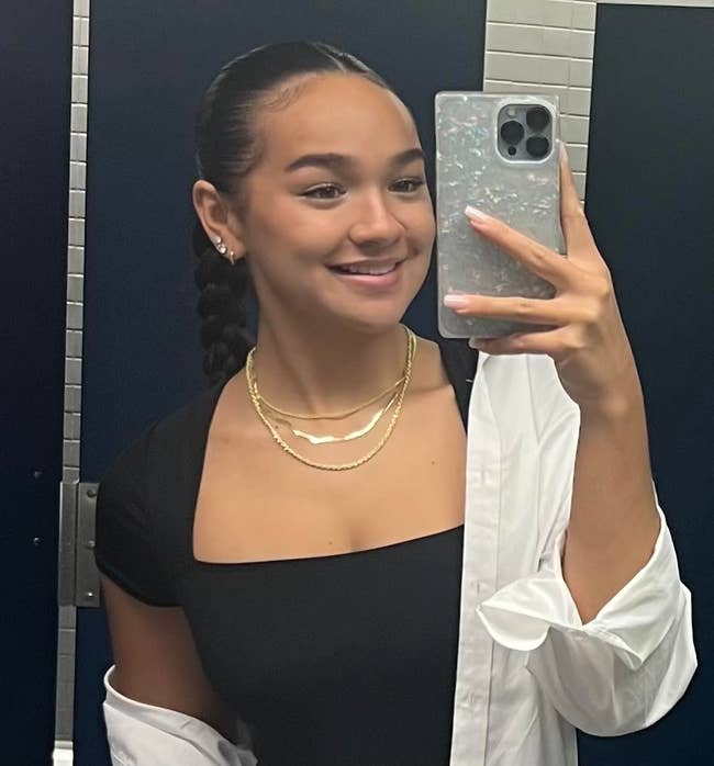 A woman takes a mirror selfie in a restroom, wearing a black top and layered necklaces, smiling as she holds a smartphone with a decorative case