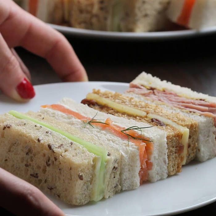 Fingers holding a plate of assorted finger sandwiches with various fillings, garnished with a sprig of dill