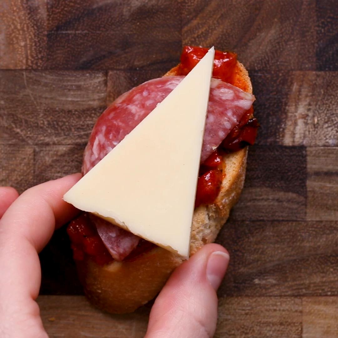 A hand holding a slice of baguette topped with chopped tomatoes, salami, and a triangular piece of cheese