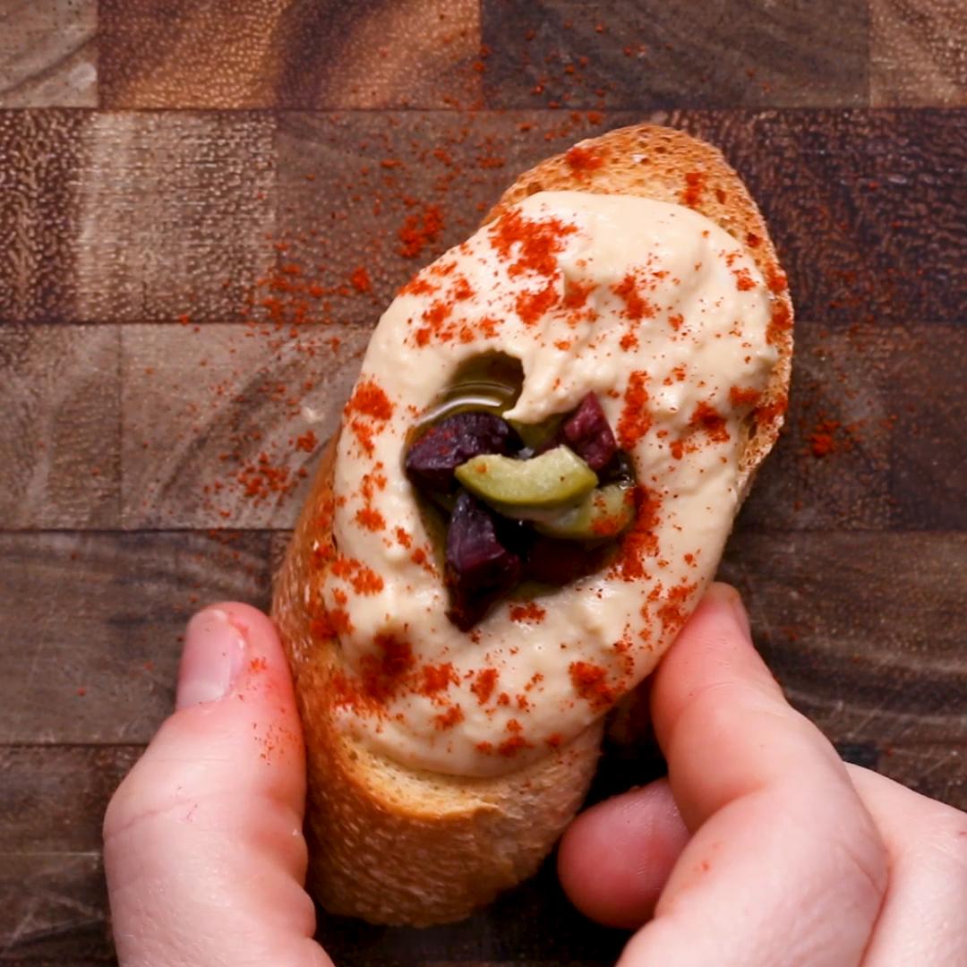 Slice of bread topped with creamy spread, olives, and paprika, held in two hands