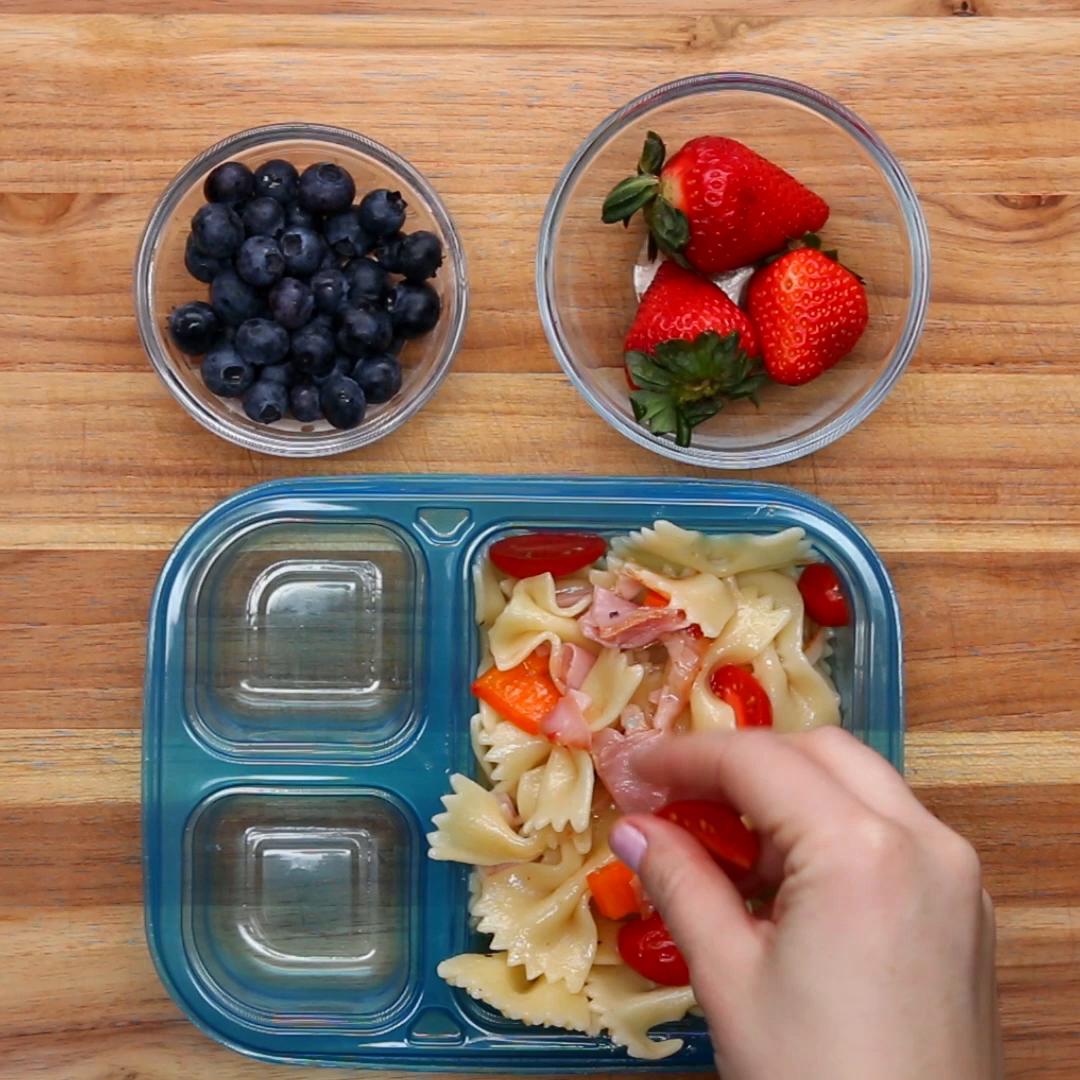 A hand prepares a lunchbox with bow tie pasta, ham, and vegetables, accompanied by bowls of blueberries and strawberries on a wooden surface