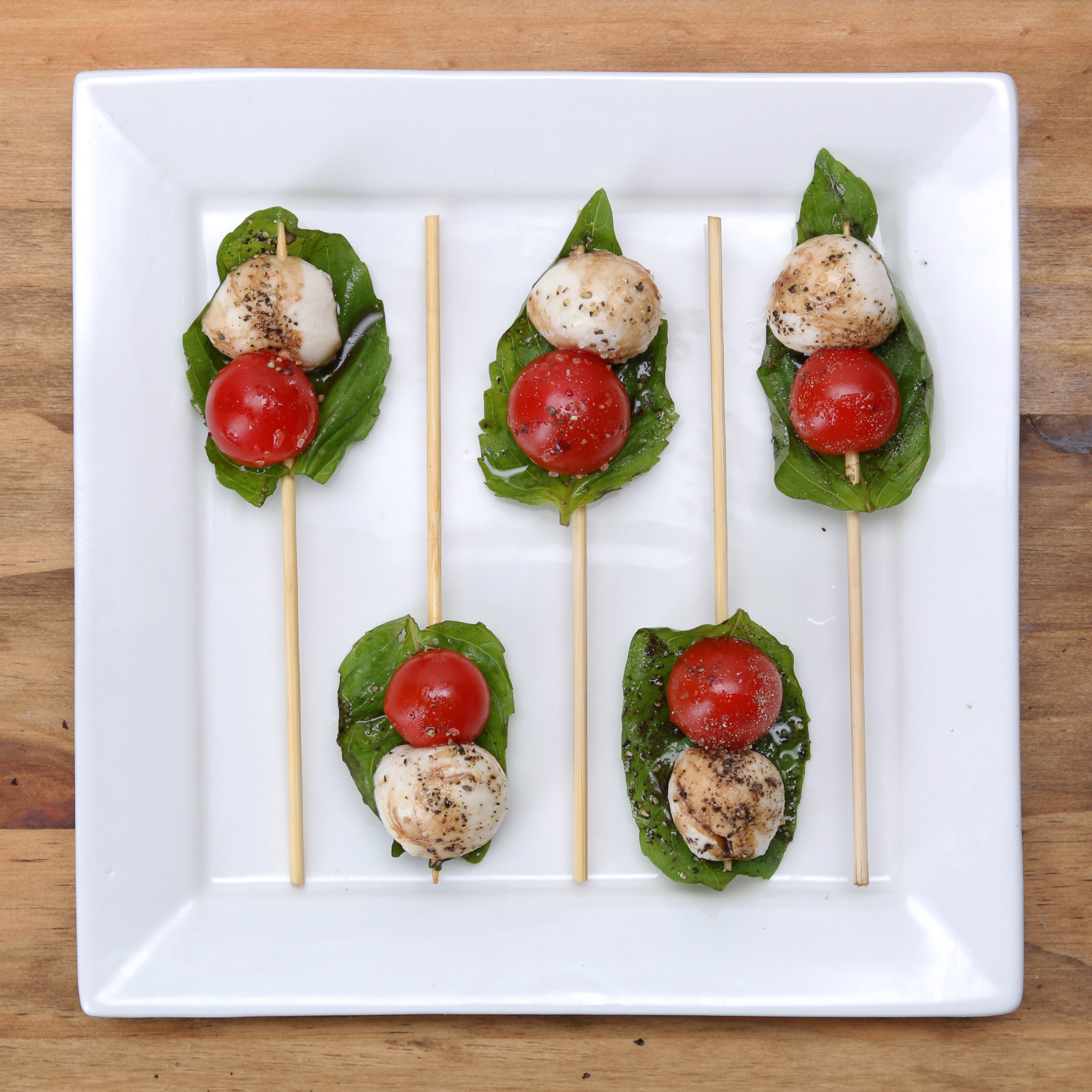 Caprese salad skewers with cherry tomatoes, mozzarella balls, and basil leaves on a white square plate