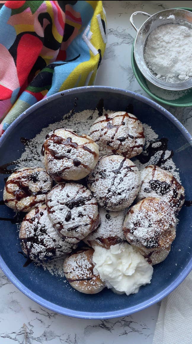 A plate of powdered sugar-dusted pastries drizzled with chocolate, served with a dollop of cream and a colorful cloth beside