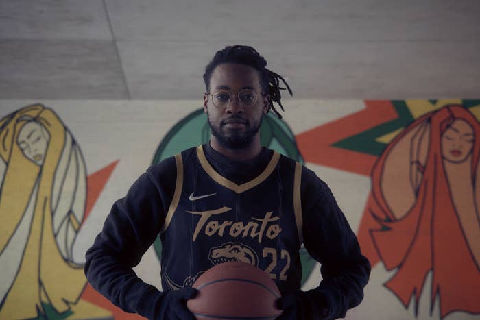 Photo of Gordon Rowe Centred, wearing a black and gold Raptors jersey and holding a basketball. Behind him is a colourful mural featuring line-drawn women.