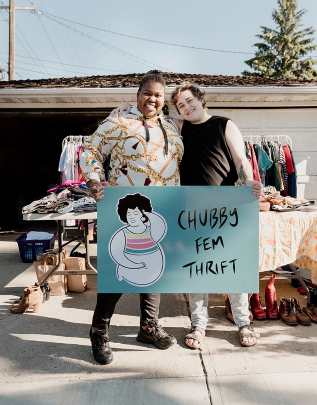 The owners of Chubby Fem Thrift holding a sign with their logo in front of their merchandise.