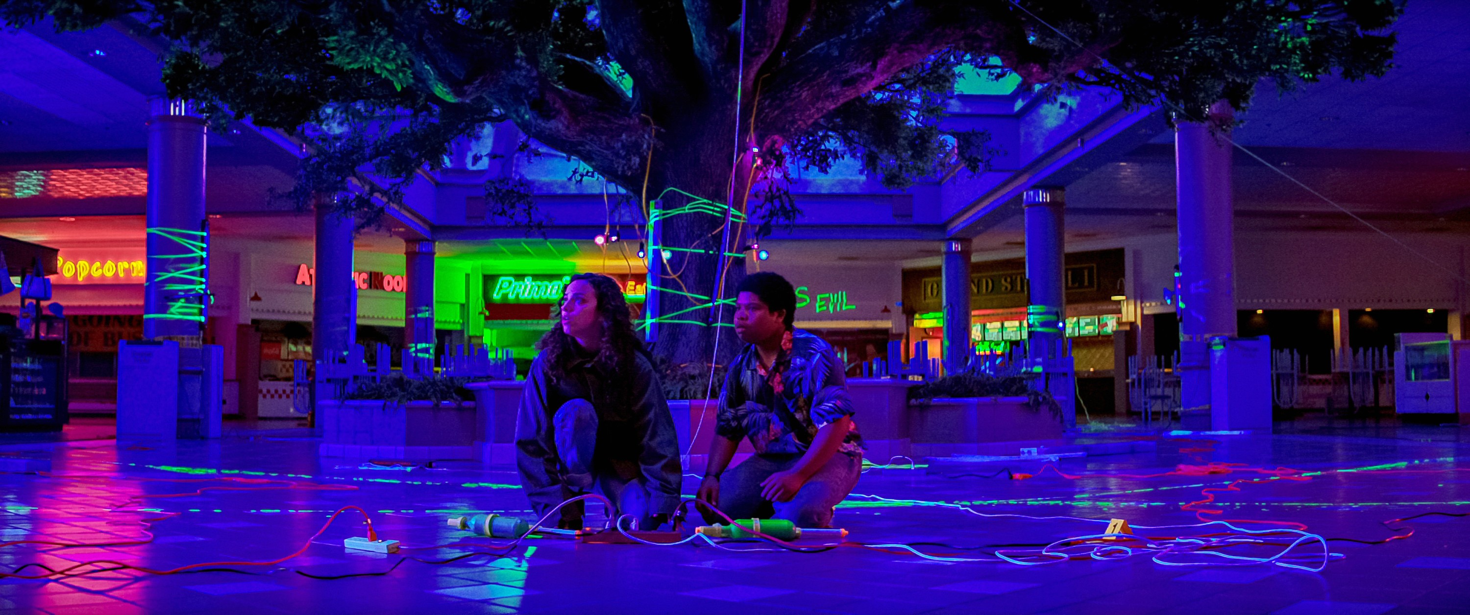 two characters sit in a neon lit mall.