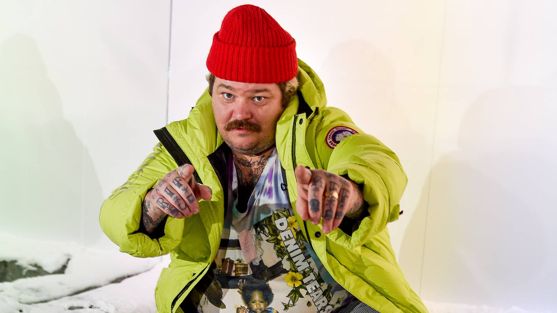 Matty Matheson wearing a read touque, a lime green Canada Goose jacket, and a graphic tee, pointing at the camera.