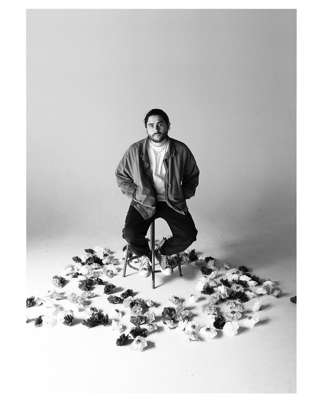 A black and white image of Ernest Carbral sitting on a stool, surrounded by flowers on the floor.