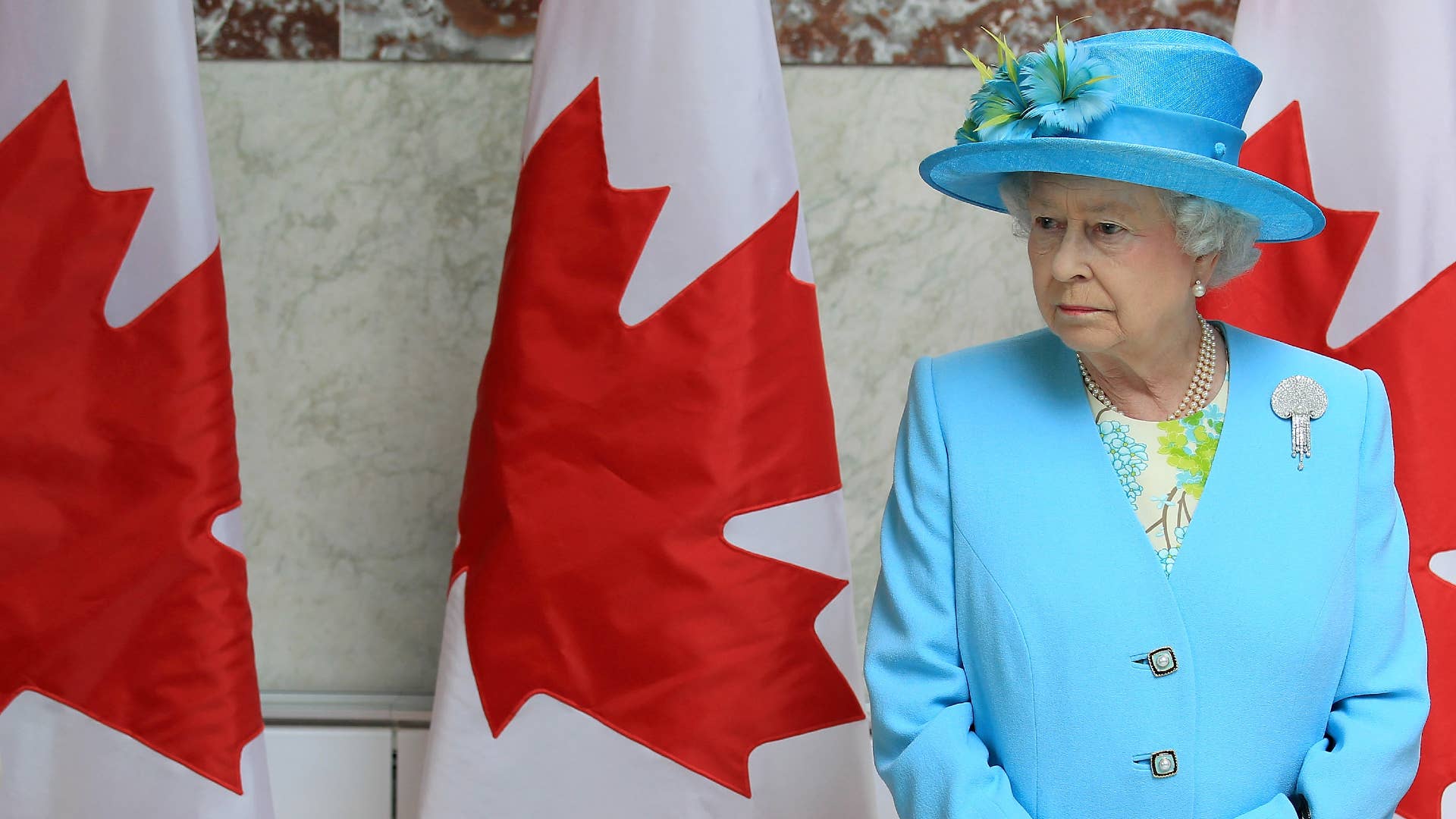 Queen Elizabeth II wearing a bright blue hat and jacket, standing in front of three Canadian flags.