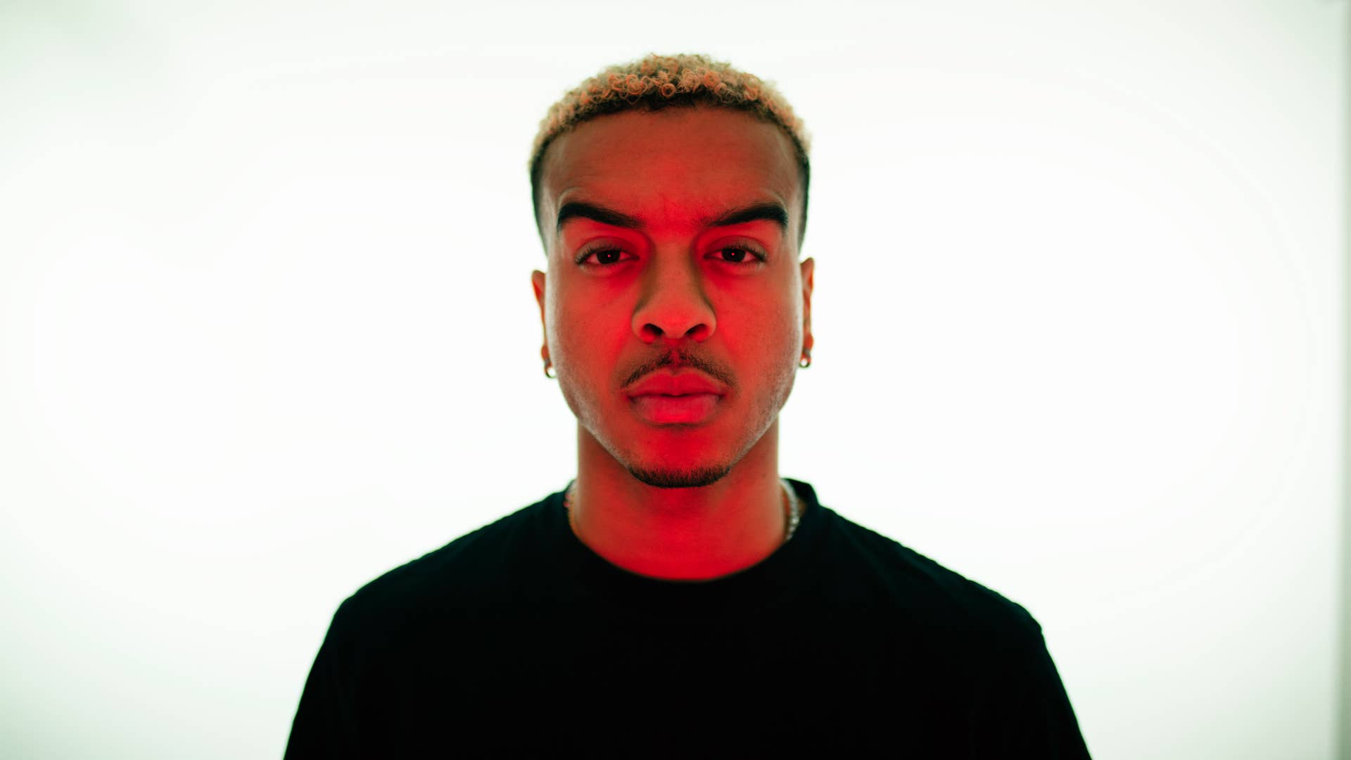 A photo of Sage Harris with red lighting on his face. He is wearing a black t-shirt against a white background.