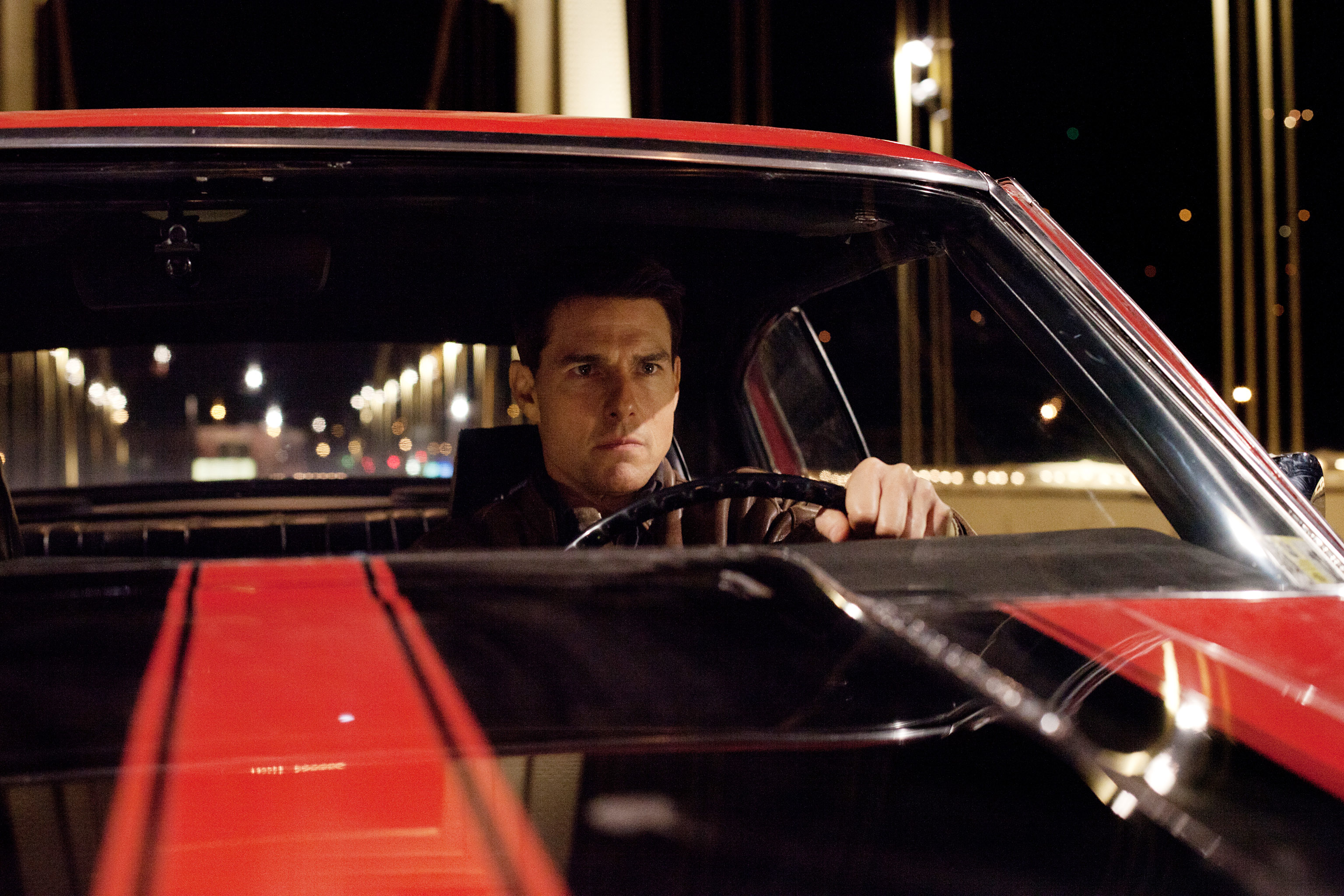 Tom Cruise sits behind the wheel of a red and black striped car.
