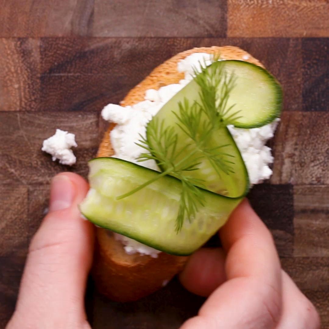 A hand holding a piece of bread topped with crumbled cheese, thin cucumber slices, and a sprig of dill