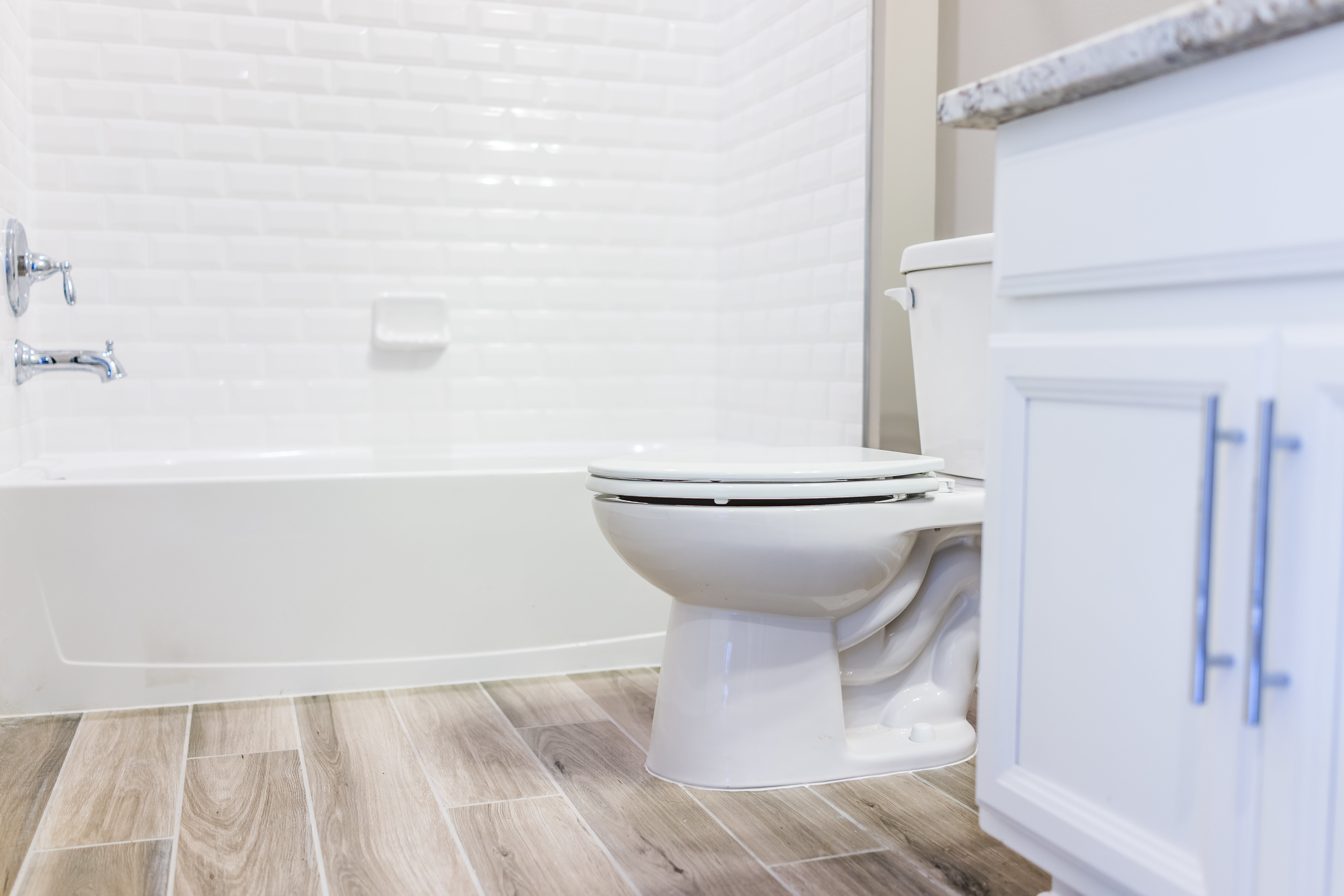 A modern bathroom with a white toilet, white bathtub with a tile backsplash, wooden floor, and a white cabinet with a granite countertop