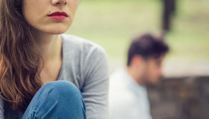 A woman in the foreground looks thoughtful, with a man sitting behind her out of focus, depicting a relationship issue