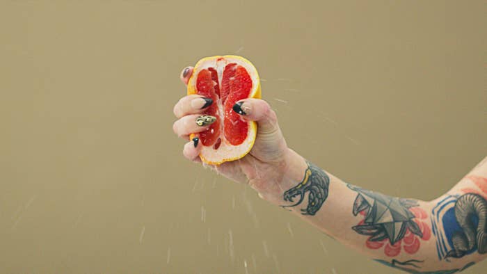 A hand with tattoos squeezes a grapefruit, juice squirting out