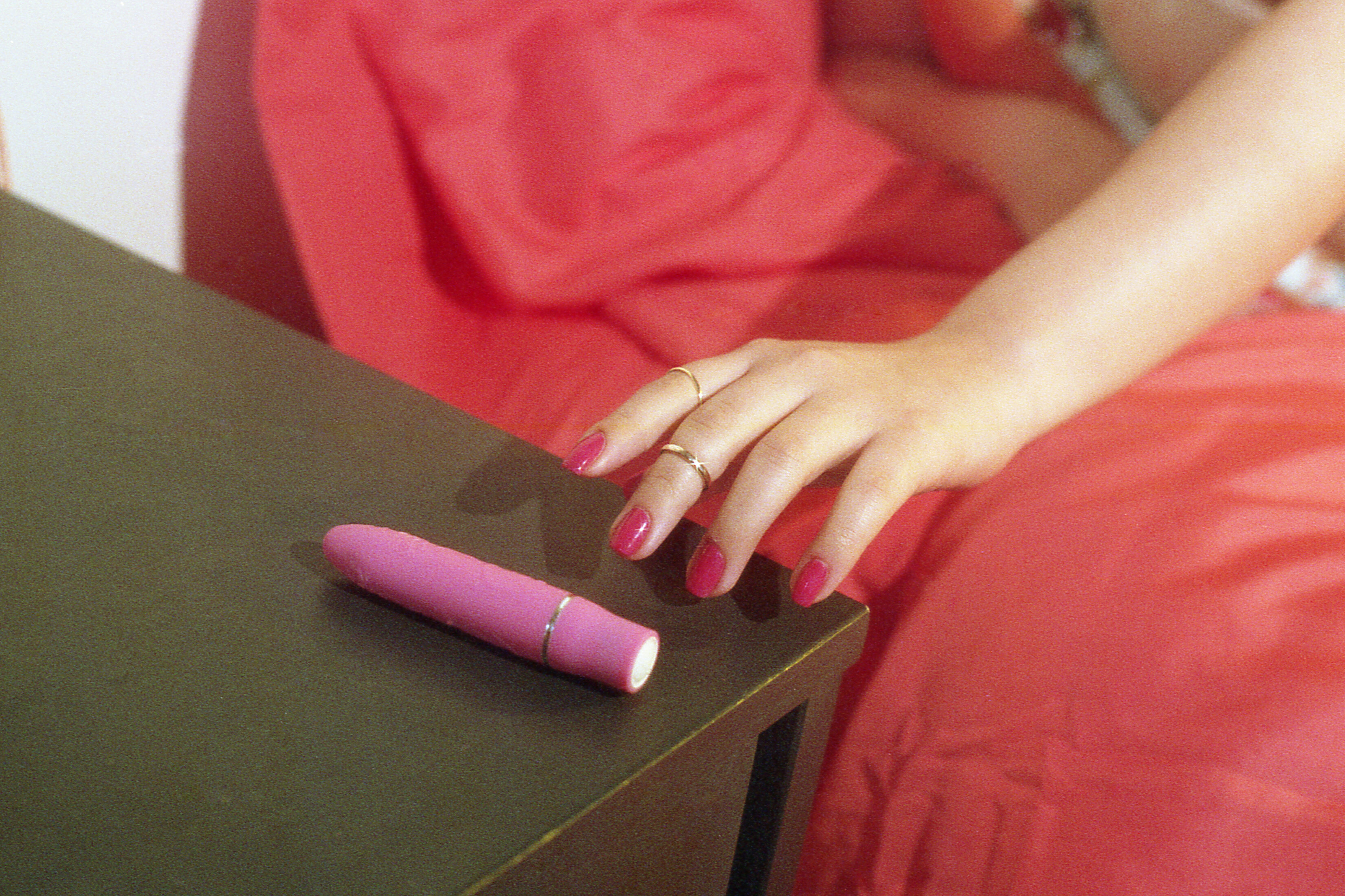 A person&#x27;s hand with rings resting next to a pink object on a dark surface, against a red fabric background