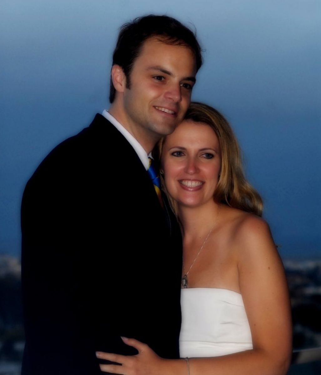 A couple dressed in formal attire embracing and smiling for the camera in an outdoor setting