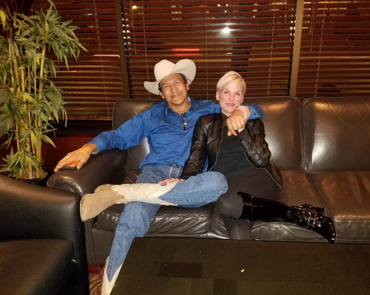 Two people sit on a couch. The person on the left wears a cowboy hat and denim outfit. The person on the right wears a leather jacket and black boots