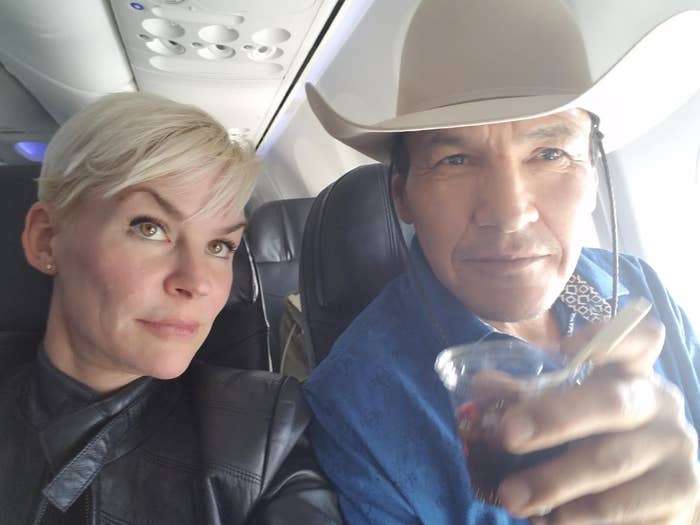 Two people sit on an airplane; one wears a leather jacket, the other a cowboy hat holding a drink