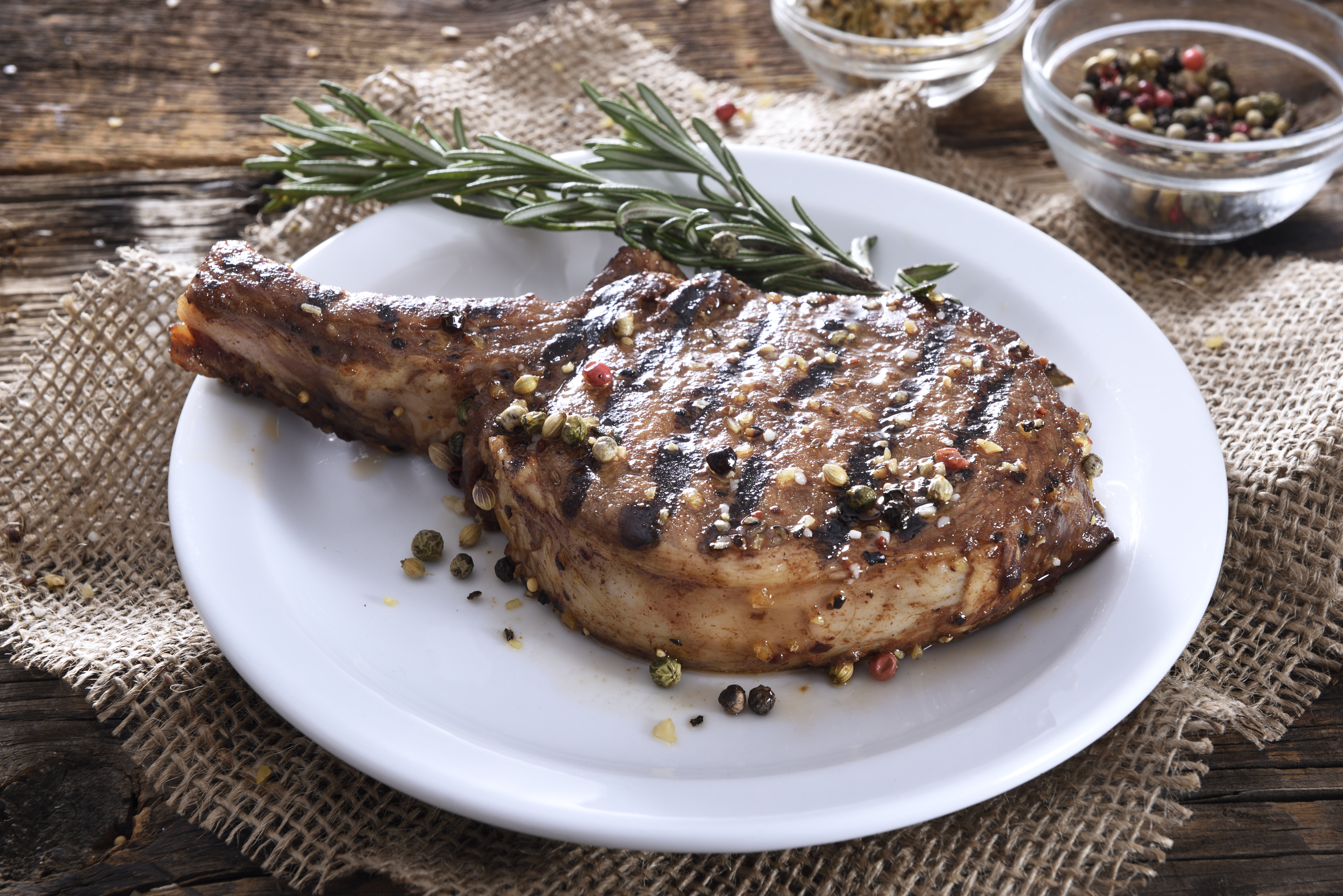 Grilled bone-in steak seasoned with pepper and herbs on a white plate, garnished with sprigs of rosemary, placed on a rustic wooden table