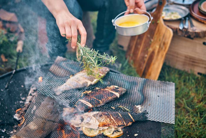 Three grilled fish on a barbecue grate being garnished with herbs by an individual's hand. Another hand holds a pot with melted butter. Plates are visible in the background