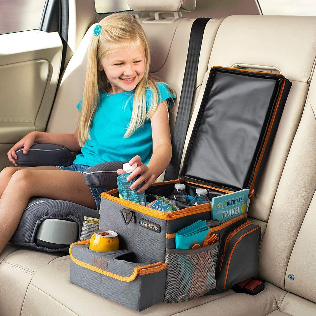 A young girl is sitting in a car seat next to a travel organizer on the backseat, which holds water bottles, snacks, and other travel essentials