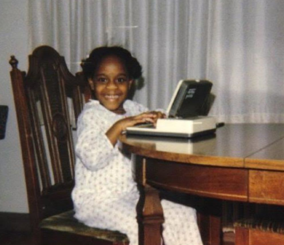 Child smiling at a table with an early laptop-style device, wearing patterned pajamas