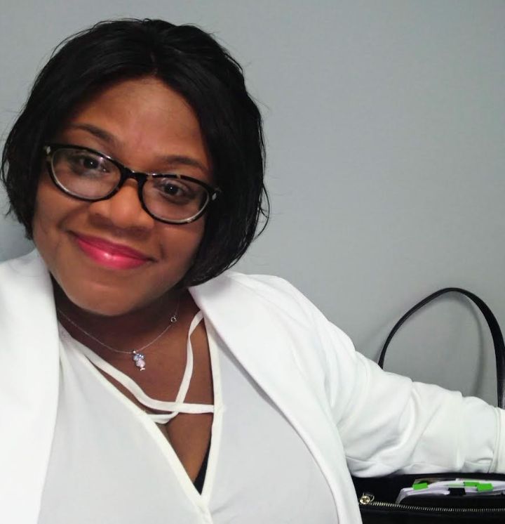 Person smiling, wearing glasses and a white blazer, sitting with a black bag beside them