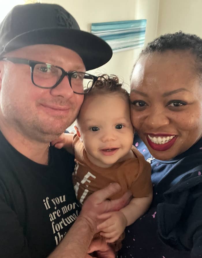 Two smiling adults holding a happy baby, posing for a cheerful family photo indoors