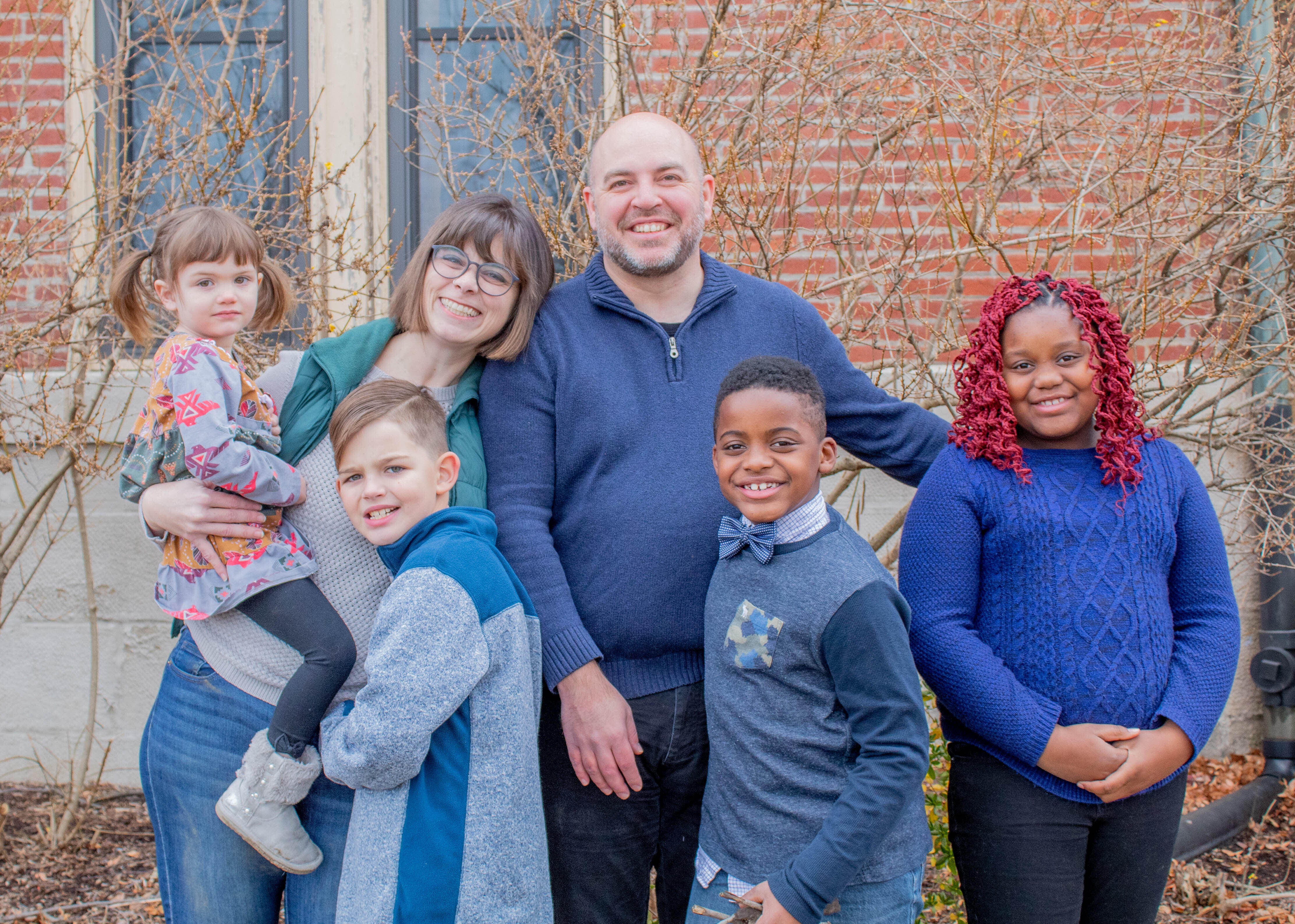 A family of six posing outdoors in casual clothing, smiling and standing close together with bare branches and a brick wall in the background