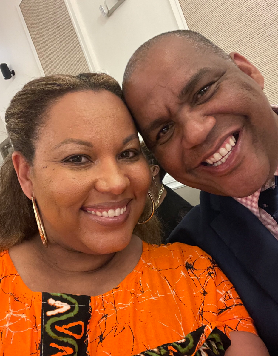 Two people smiling closely together for a photo, with one wearing a patterned top and the other in a suit