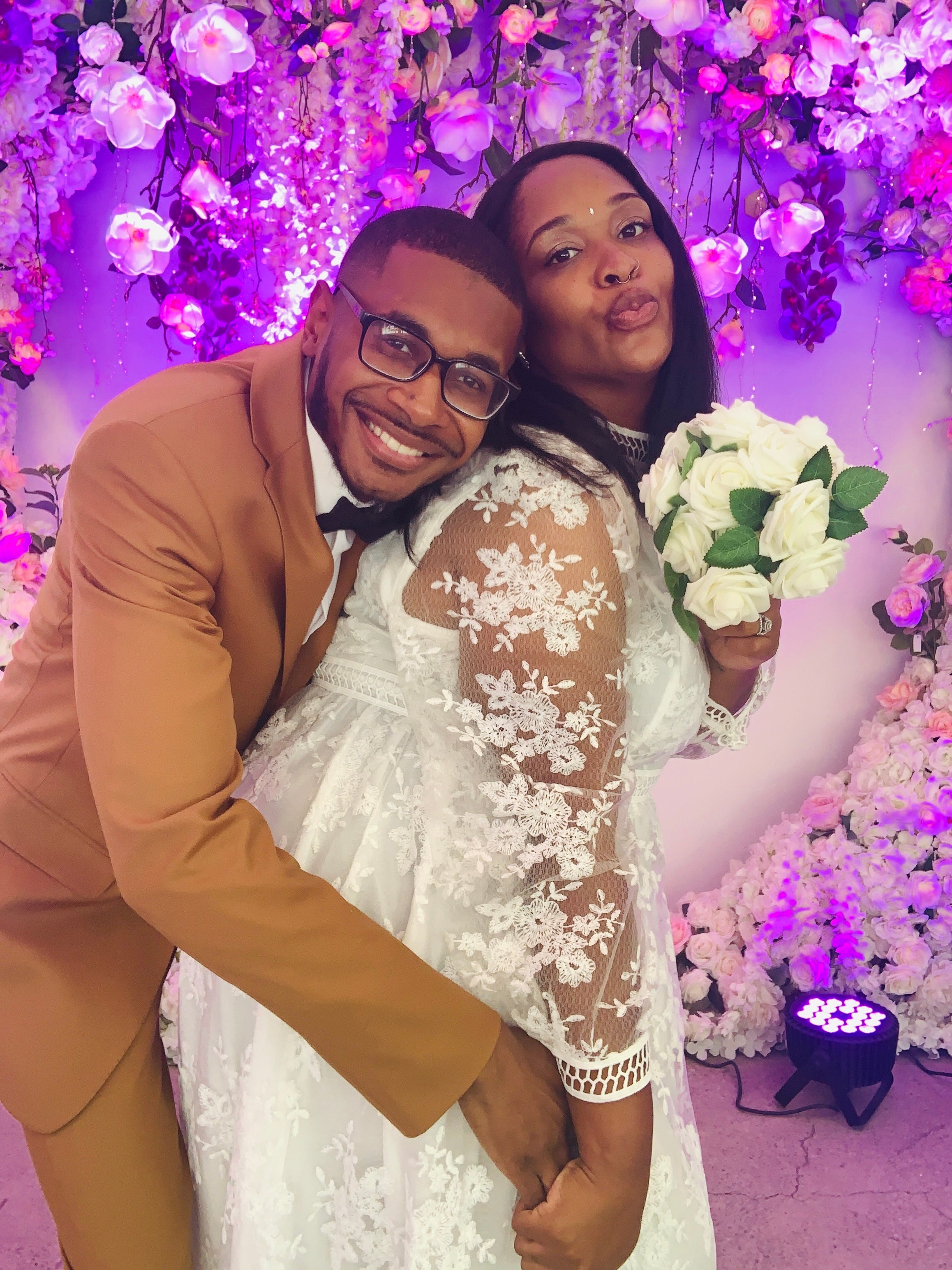 A joyful couple at a special event. The man in a brown suit hugs the woman who is wearing a lace dress and holding a bouquet of white roses