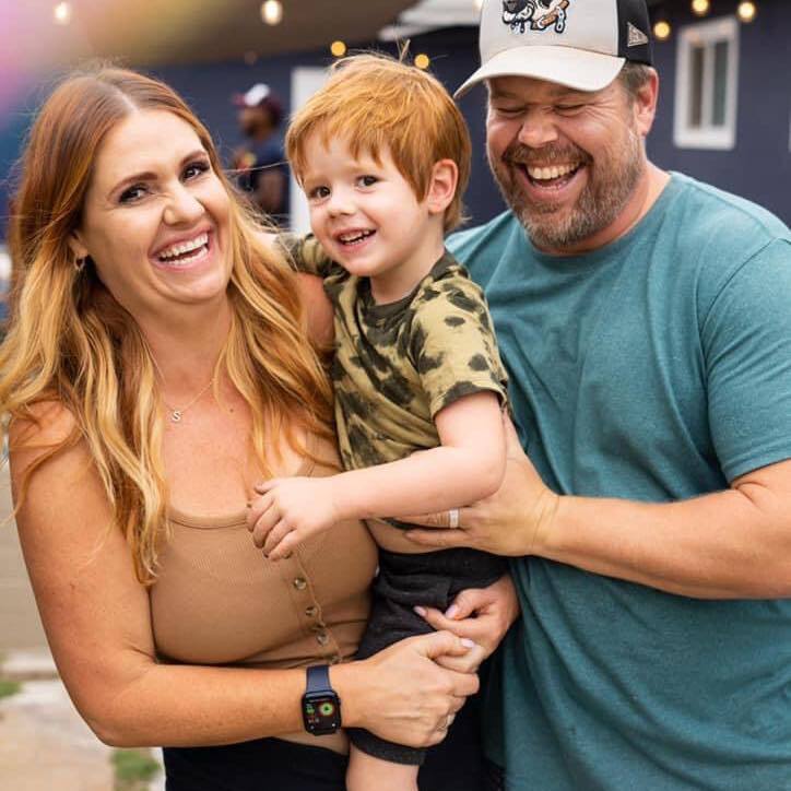 Smiling family of three poses outdoors. The woman is in a sleeveless top, the man in a cap and t-shirt, holding a young boy in a camo shirt