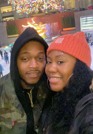 A couple smiles for a selfie in front of a bustling city ice rink with a large illuminated sculpture in the background
