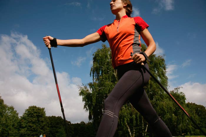 A person in athletic attire is walking with hiking poles in an outdoor setting with trees and a cloudy sky in the background