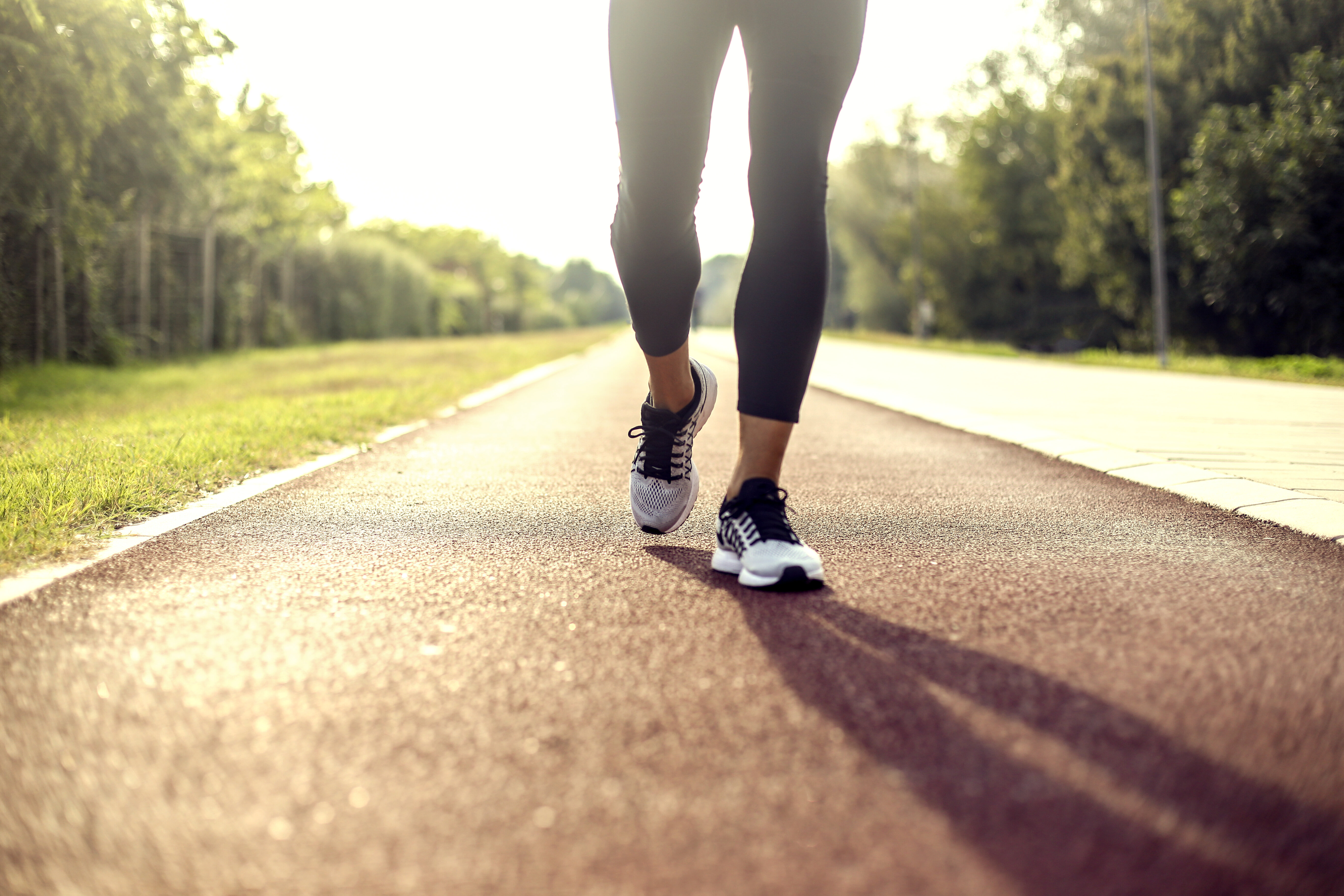 A person walking on a paved path in an outdoor setting, wearing athletic shoes and leggings. The path is surrounded by trees and greenery