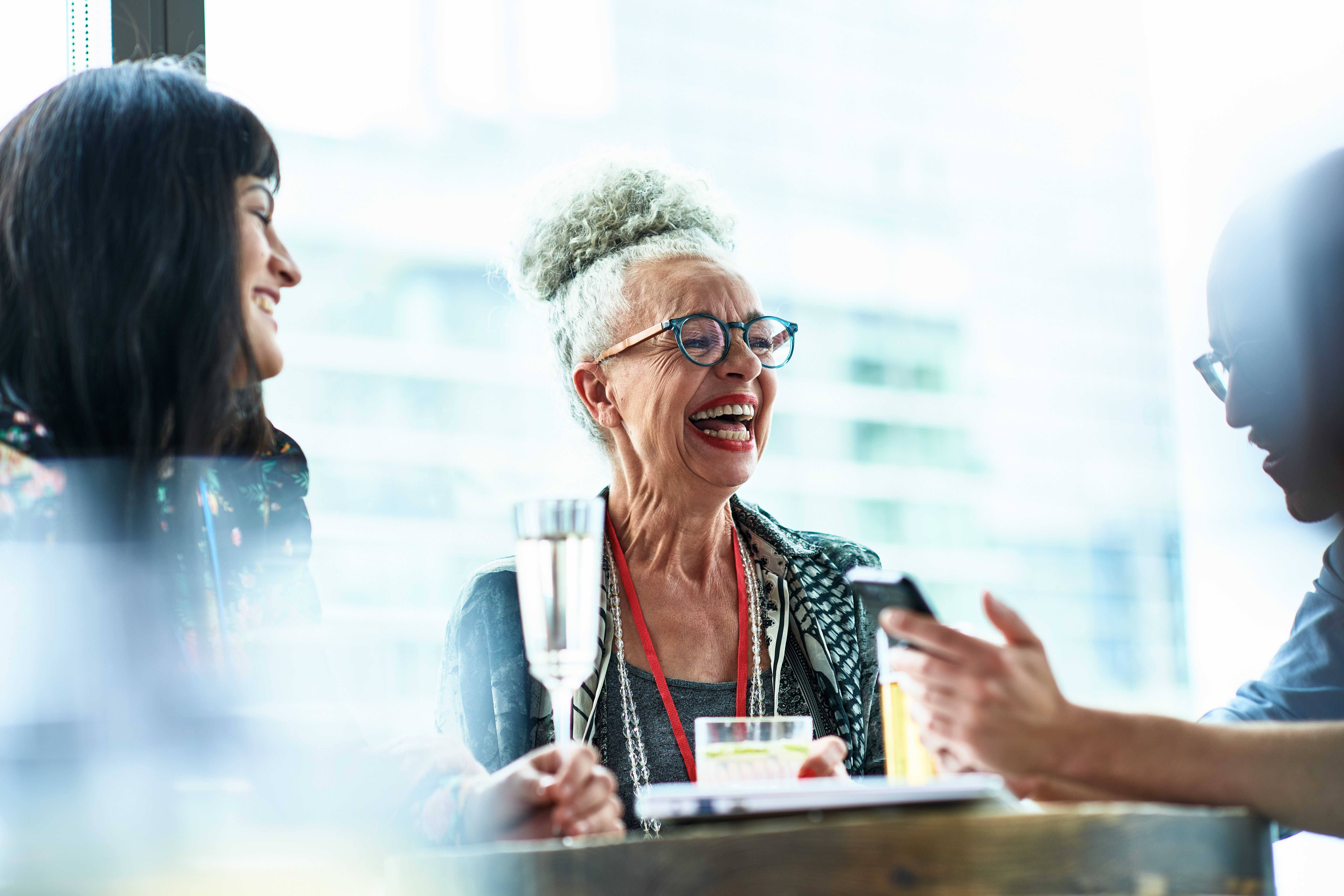 A group of people, including an elderly woman with glasses and gray hair, are laughing and enjoying drinks together at a table