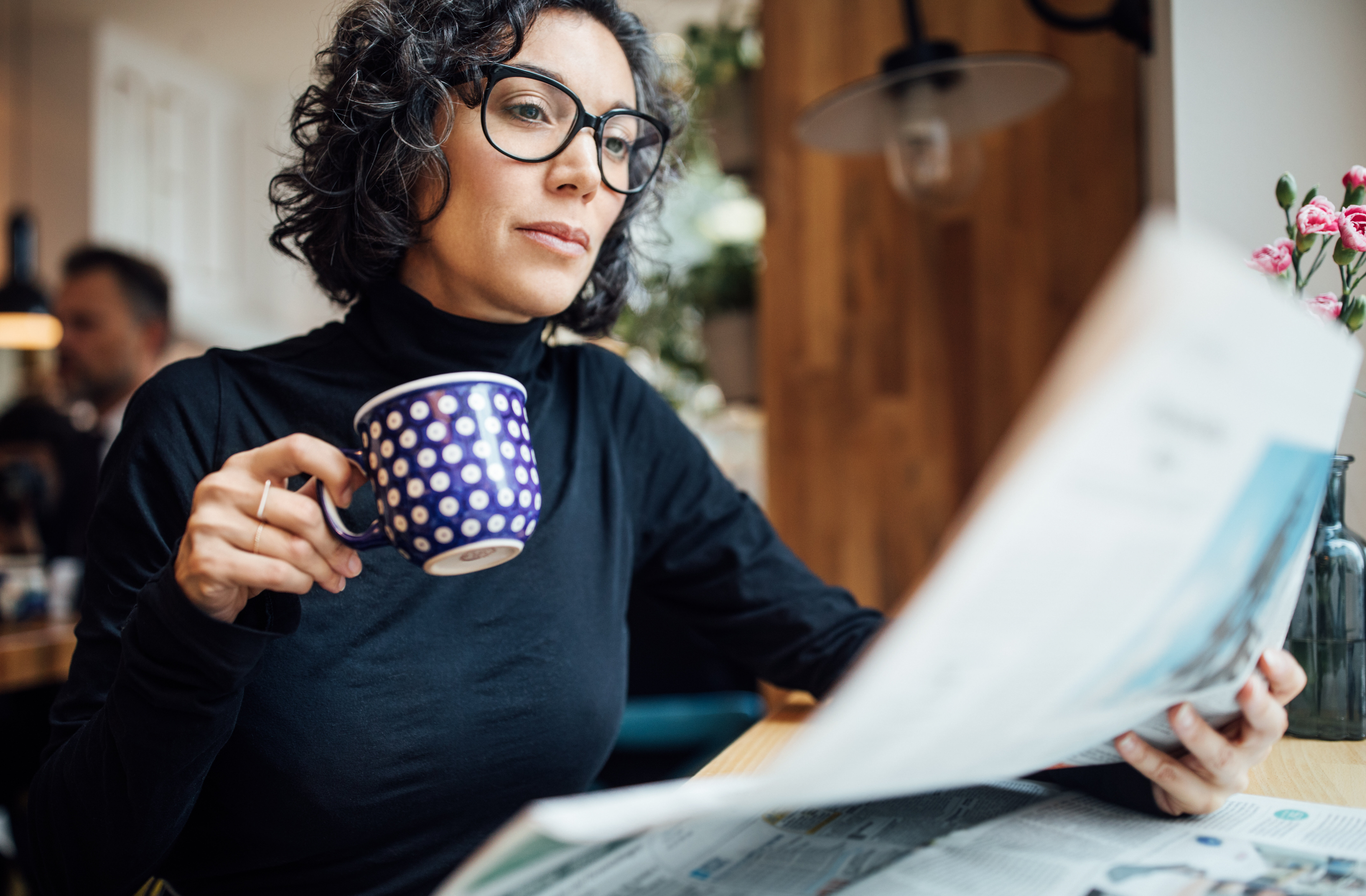 A person with curly hair and glasses reads a newspaper while holding a polka-dot mug in a cozy indoor setting with flowers in the background